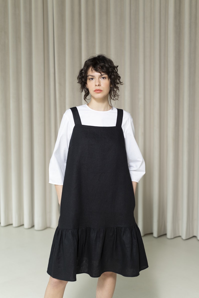 K&ouml;nnte beinhalten: Ein schwarzes Leinenkleid mit R&uuml;schen am Saum und d&uuml;nnen Tr&auml;gern. Das Kleid wird &uuml;ber einem wei&szlig;en T-Shirt getragen.