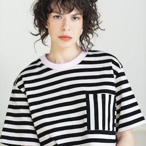 May include: A black and white striped t-shirt with a pocket. The t-shirt has a pink collar and short sleeves. The model is wearing the shirt and has dark curly hair.