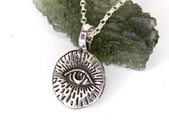 Puede incluir: Collar de plata con un colgante redondo que presenta un ojo con líneas radiantes. El colgante descansa sobre un cristal verde.