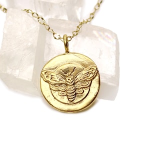 May include: A gold necklace with a round pendant featuring a detailed embossed image of a moth.