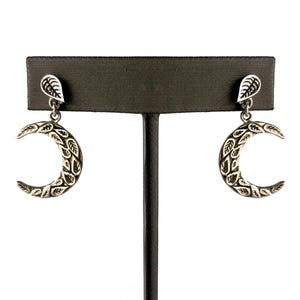 Pode incluir: Brincos de lua crescente prateados com detalhes de folhas. Cada brinco apresenta um formato de lua crescente com intrincados padrões de folhas e um pequeno pino em forma de folha. Os brincos são exibidos em um suporte de joias preto sobre um fundo branco.
