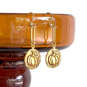 May include: Gold-colored pumpkin earrings. Each earring features a small, oval-shaped charm with a detailed pumpkin design. The earrings hang from a gold-colored hook and chain. The background is a blurred amber-colored glass.