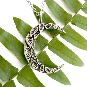May include: A silver crescent moon pendant necklace with a detailed leaf design. The pendant is hanging on a silver chain and is resting on a green fern leaf.
