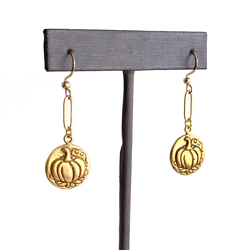May include: Gold-colored dangle earrings. Each earring features a circular charm with a pumpkin design. The pumpkin is surrounded by a decorative border. The earrings hang from a gold-colored chain and hook.