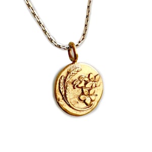 May include: A gold-colored pendant necklace featuring a circular charm with a floral and wheat design. The pendant hangs from a delicate silver chain, creating a classic and elegant look. The charm's details are finely etched, showcasing the intricate floral pattern.