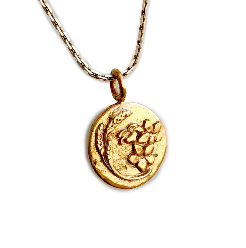 May include: A gold-colored pendant necklace featuring a circular charm with a floral and wheat design. The pendant hangs from a delicate silver chain, creating a classic and elegant look. The charm's details are finely etched, showcasing the intricate floral pattern.
