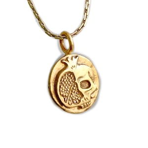 May include: Gold-toned pendant necklace featuring a detailed design of a pomegranate and a skull. The pendant is round and hangs from a delicate chain. The design is a combination of a pomegranate and a skull, creating a unique and symbolic piece.