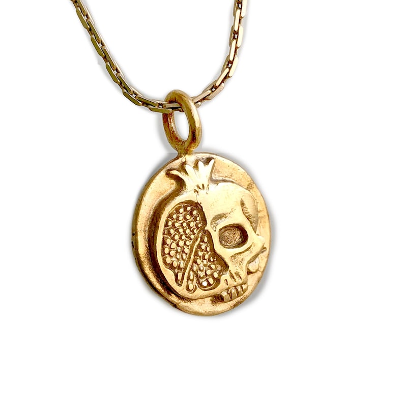 May include: Gold-toned pendant necklace featuring a detailed design of a pomegranate and a skull. The pendant is round and hangs from a delicate chain. The design is a combination of a pomegranate and a skull, creating a unique and symbolic piece.