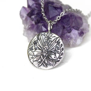 May include: A silver necklace with a round pendant featuring an embossed design of a fairy with wings spread, surrounded by flowers and leaves.