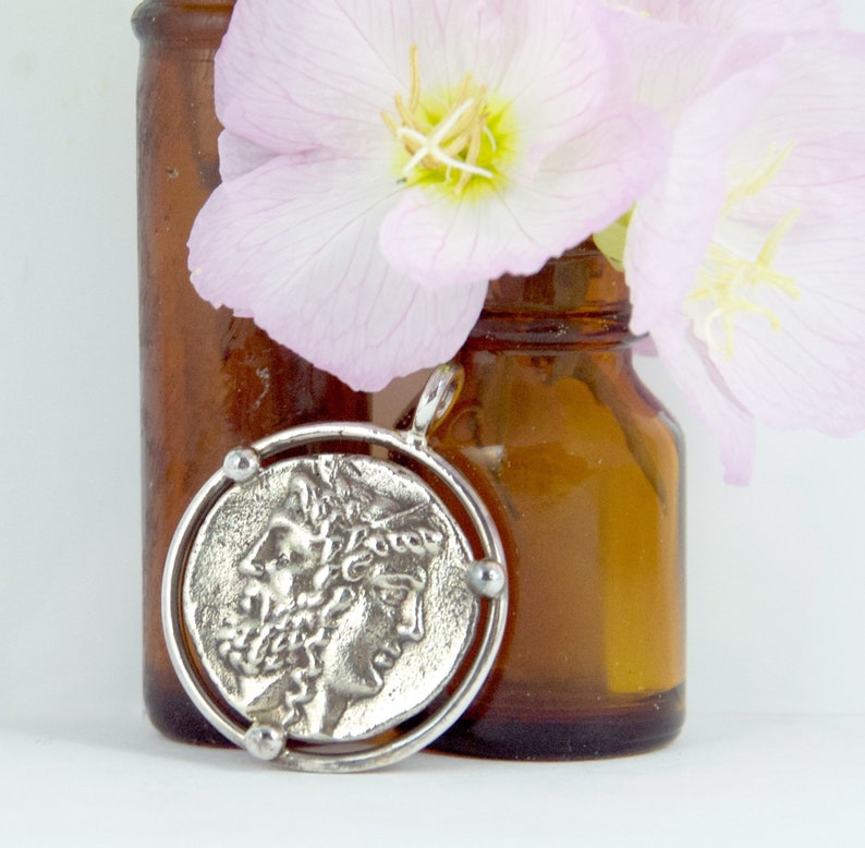 May include: A silver pendant featuring a round coin with a profile of a woman's face. The coin is surrounded by a silver ring with small decorative balls on the sides.