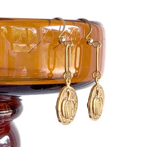 May include: Gold-colored earrings with a pumpkin design. The earrings have a hook closure and a long, rectangular link. The pumpkin pendant is oval-shaped with a detailed carving of a pumpkin.