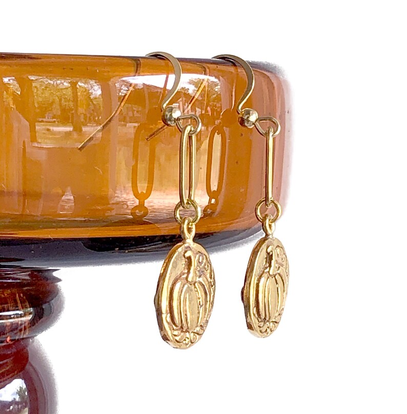 May include: Gold-colored earrings with a pumpkin design. The earrings have a hook closure and a long, rectangular link. The pumpkin pendant is oval-shaped with a detailed carving of a pumpkin.
