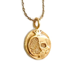 May include: A gold-colored pendant necklace featuring a unique design. The pendant is oval-shaped and depicts a pomegranate combined with a skull. The necklace has a delicate chain and is set against a white background.