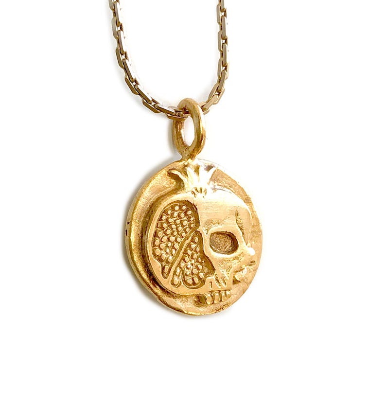 May include: A gold-colored pendant necklace featuring a unique design. The pendant is oval-shaped and depicts a pomegranate combined with a skull. The necklace has a delicate chain and is set against a white background.