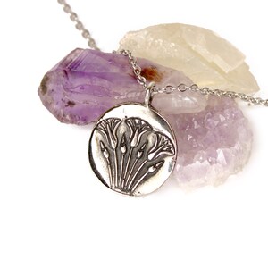 May include: A silver necklace with a round pendant featuring an embossed design of stylized flowers. The pendant is resting on a cluster of amethyst and clear quartz crystals.