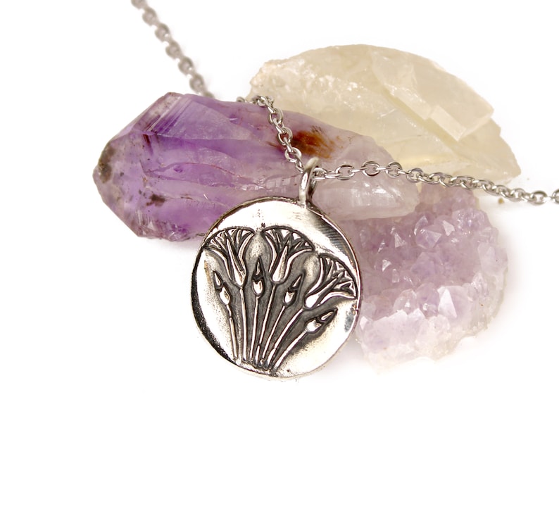May include: A silver necklace with a round pendant featuring an embossed design of stylized flowers. The pendant is resting on a cluster of amethyst and clear quartz crystals.