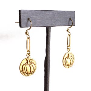 May include: Gold-colored dangle earrings. Each earring features a small, circular charm with a pumpkin design. The charms are suspended from a gold chain and hook. The earrings are displayed on a gray stand against a white background.