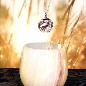 May include: A silver chain necklace with a round pendant featuring a detailed image of a bird. The pendant is hanging above a white alabaster bowl with pink streaks.