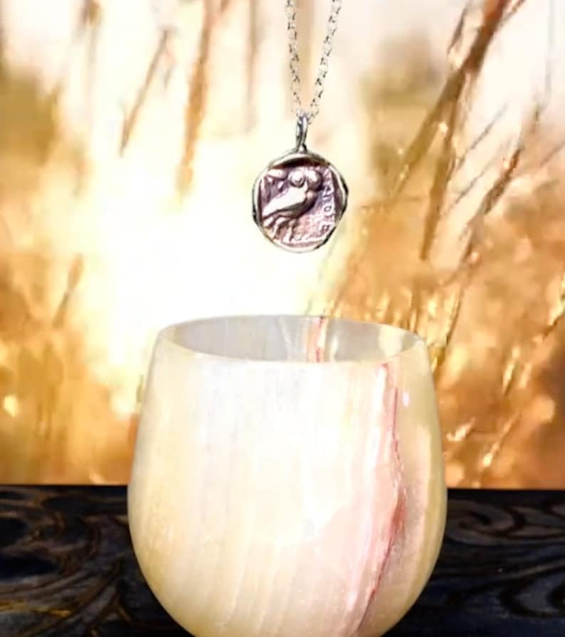 May include: A silver chain necklace with a round pendant featuring a detailed image of a bird. The pendant is hanging above a white alabaster bowl with pink streaks.