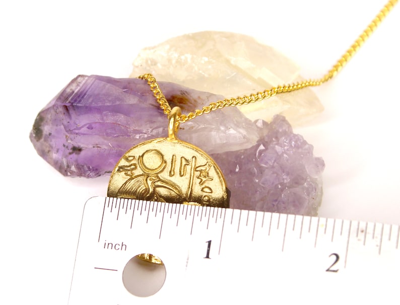 May include: Gold necklace with a round pendant featuring an Egyptian hieroglyphic design. The pendant is about 1 inch in diameter and is resting on a purple amethyst crystal.