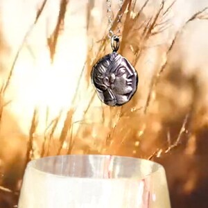 May include: A silver necklace with a pendant featuring a detailed profile of a woman's face. The pendant is round and has a textured surface. The necklace is hanging against a backdrop of dried grass.