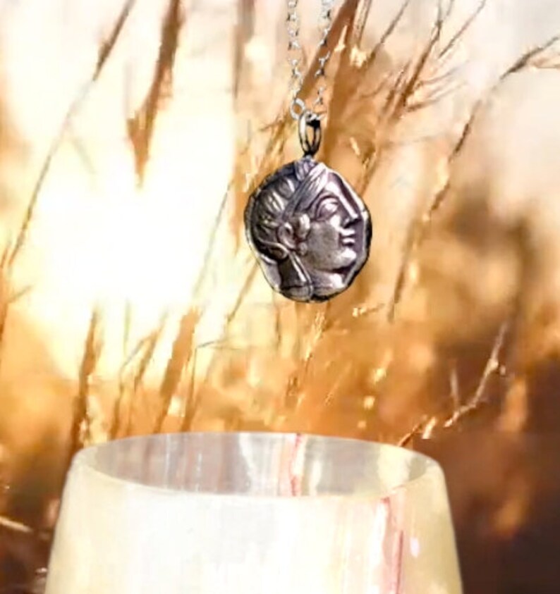 May include: A silver necklace with a pendant featuring a detailed profile of a woman's face. The pendant is round and has a textured surface. The necklace is hanging against a backdrop of dried grass.