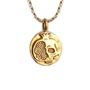 May include: Gold-toned pendant necklace featuring a detailed design of a pomegranate and a skull. The pendant is round and hangs from a delicate chain. The necklace is set against a white background.