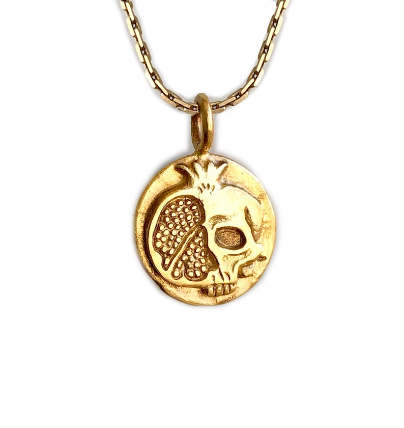 May include: Gold-toned pendant necklace featuring a detailed design of a pomegranate and a skull. The pendant is round and hangs from a delicate chain. The necklace is set against a white background.