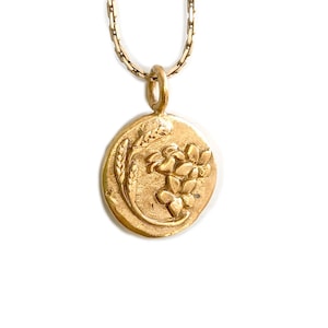 May include: Gold-toned pendant necklace featuring a circular charm with a wheat stalk and floral design. The pendant hangs from a delicate chain. The necklace is set against a white background.