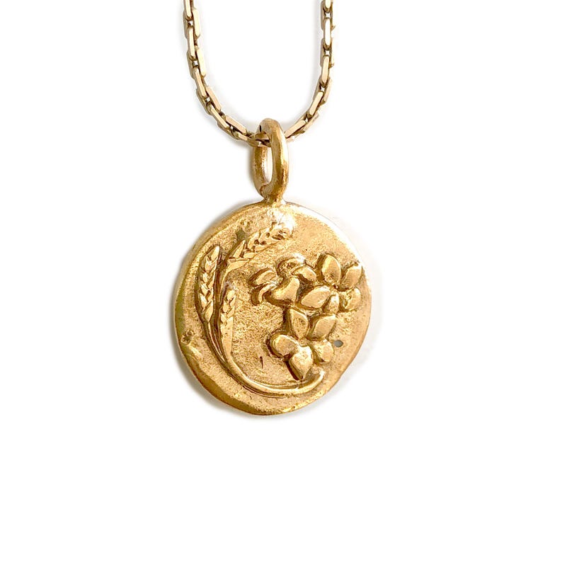 May include: Gold-toned pendant necklace featuring a circular charm with a wheat stalk and floral design. The pendant hangs from a delicate chain. The necklace is set against a white background.