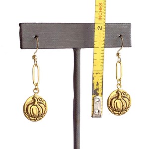 May include: Gold-colored earrings with a pumpkin design. The earrings are approximately 2 inches long. The pumpkin is in the center of a circular pendant with a decorative border. The earrings hang from a gold-colored hook.