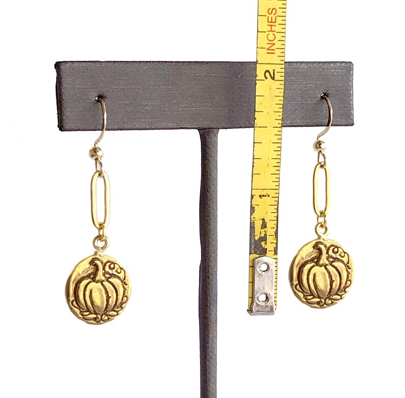 May include: Gold-colored earrings with a pumpkin design. The earrings are approximately 2 inches long. The pumpkin is in the center of a circular pendant with a decorative border. The earrings hang from a gold-colored hook.