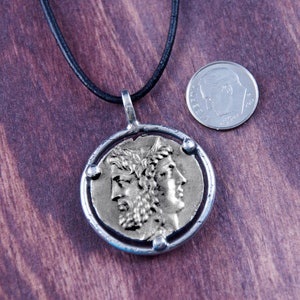 May include: Silver pendant necklace with a round coin featuring a profile of a person with a crown. The coin is surrounded by a silver ring with three small silver dots. The necklace is on a black leather cord.