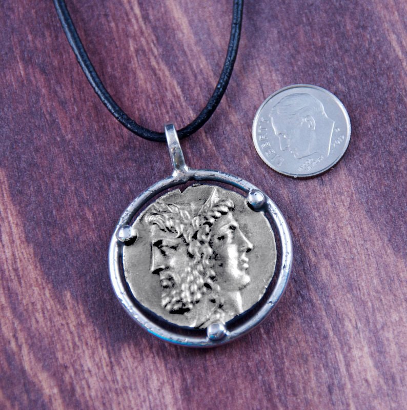 May include: Silver pendant necklace with a round coin featuring a profile of a person with a crown. The coin is surrounded by a silver ring with three small silver dots. The necklace is on a black leather cord.