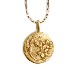 May include: A gold-colored pendant necklace featuring a circular charm with a floral and wheat design. The pendant hangs from a delicate chain. The charm has a textured, antique finish, and the design is raised.