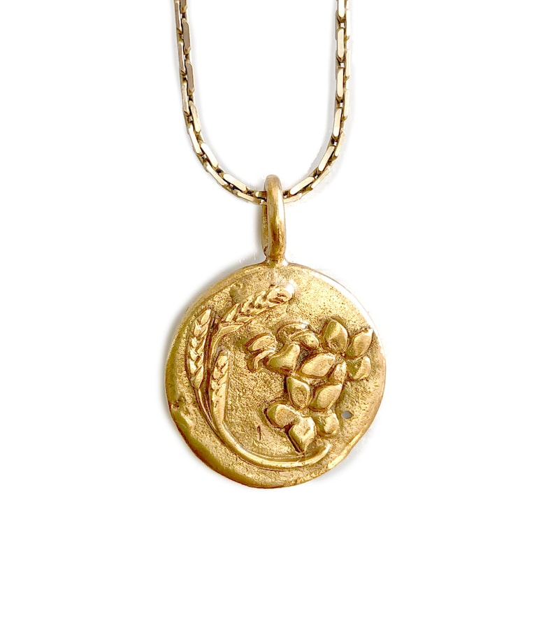 May include: A gold-colored pendant necklace featuring a circular charm with a floral and wheat design. The pendant hangs from a delicate chain. The charm has a textured, antique finish, and the design is raised.