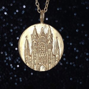 May include: A gold pendant necklace featuring a detailed embossed design of a cathedral with a large stained glass window.