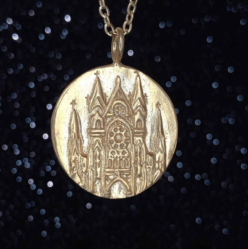May include: A gold pendant necklace featuring a detailed embossed design of a cathedral with a large stained glass window.