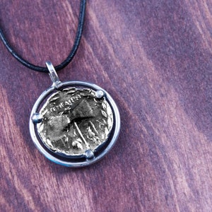 May include: A silver necklace with a round pendant featuring a gold coin with an inscription. The coin is set in a silver frame with four small silver beads.