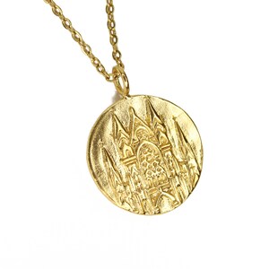 May include: Gold necklace with a round pendant featuring a detailed embossed design of a cathedral.