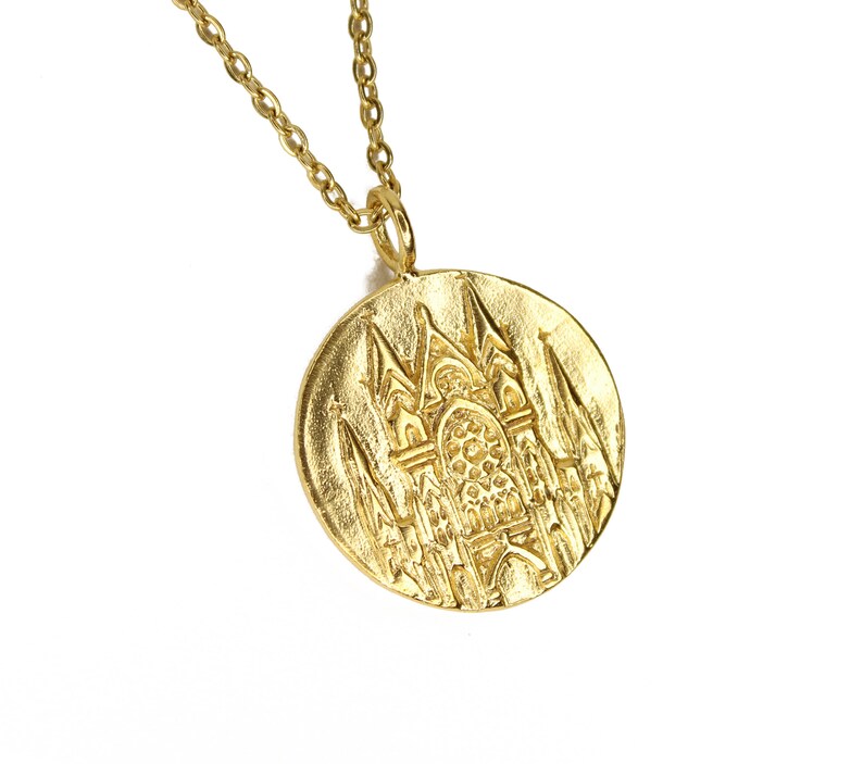 May include: Gold necklace with a round pendant featuring a detailed embossed design of a cathedral.