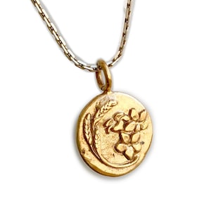 May include: Gold-toned pendant necklace featuring an oval pendant with a floral and wheat design. The pendant hangs from a delicate chain. The necklace is set against a white background.