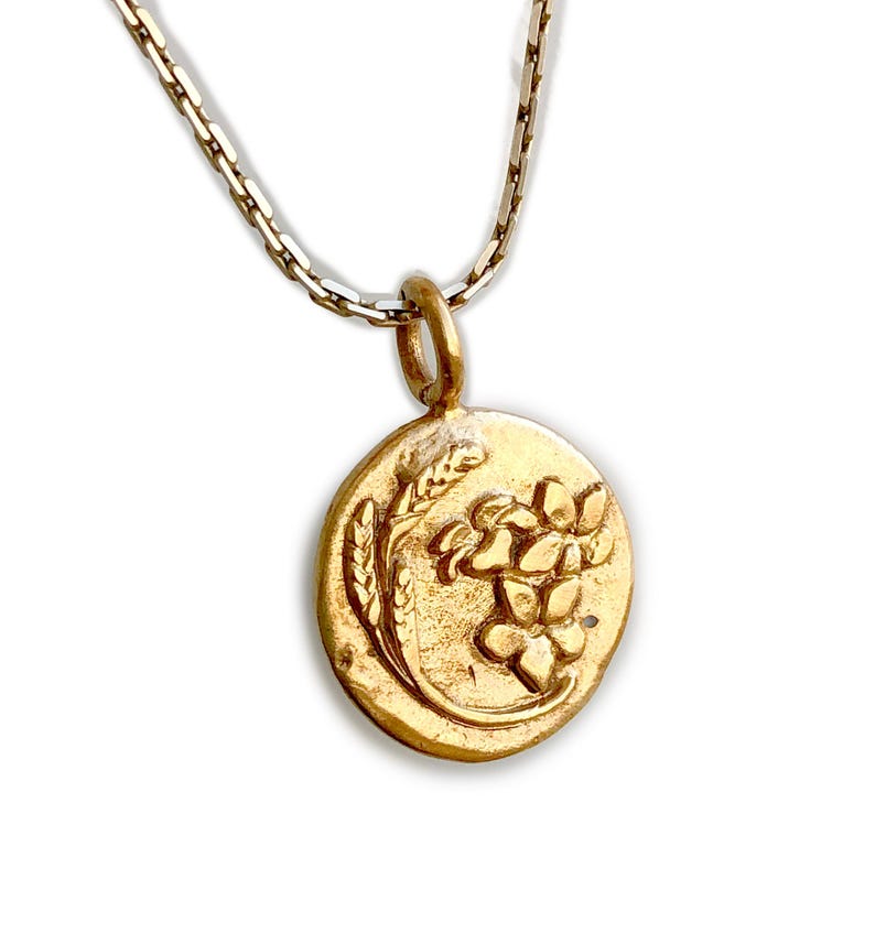 May include: Gold-toned pendant necklace featuring an oval pendant with a floral and wheat design. The pendant hangs from a delicate chain. The necklace is set against a white background.
