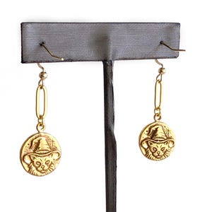 May include: Gold-colored earrings featuring a cat wearing a witch hat. The earrings have a circular pendant with the cat design and a long, rectangular link. The earrings are displayed on a gray earring stand against a white background.