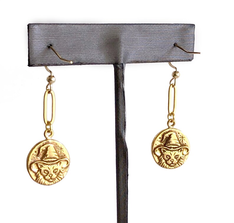 May include: Gold-colored earrings featuring a cat wearing a witch hat. The earrings have a circular pendant with the cat design and a long, rectangular link. The earrings are displayed on a gray earring stand against a white background.
