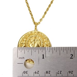 May include: Gold pendant necklace with a round, detailed coin-shaped charm. The charm is approximately 1 inch in diameter. The necklace is shown next to a ruler for size reference.