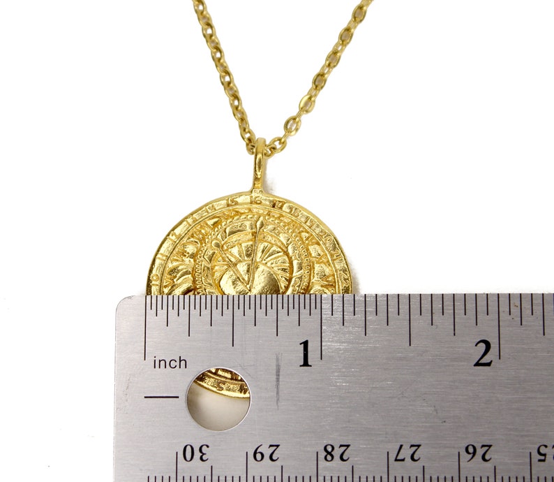 May include: Gold pendant necklace with a round, detailed coin-shaped charm. The charm is approximately 1 inch in diameter. The necklace is shown next to a ruler for size reference.