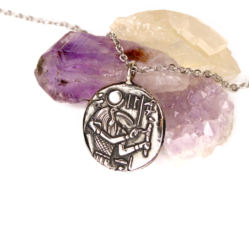May include: Silver necklace with a round pendant featuring an engraved image of an Egyptian god with a falcon head. The pendant is resting on a bed of amethyst crystals.