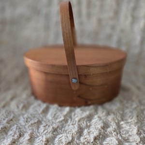 May include: A round, wooden basket with a lid and a curved handle. The basket is made of light brown wood and has a simple, classic design. The handle is attached with a small metal rivet. The basket is suitable for storage or display.