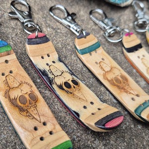 Könnte beinhalten: Miniatur-Skateboard-Schlüsselanhänger mit einem in das Holz geätzten Totenkopf-Design. Jeder Schlüsselanhänger hat ein anderes Farbschema an den Enden, darunter Grün, Blau, Rosa und Orange. Sie haben silberfarbene Verschlüsse.